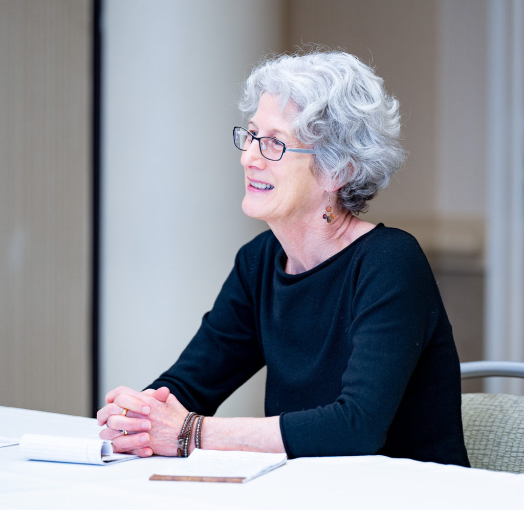 Headshot of author sitting at a conference table. She has gray hair and is wearing glasses and a long sleeved black shirt. She is smiling.