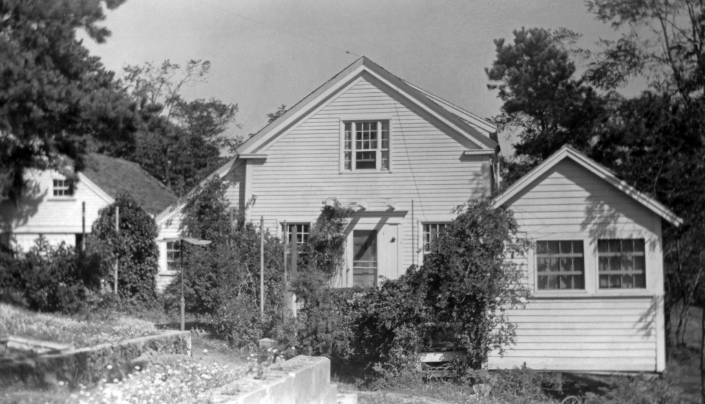 Black and white photograph of author’s home in Wellfleet in the 1950s, taken from the side yard. Ivy covers the clapboard walls. A small summerhouse stands to the right of the house, and the garage stands to the left.